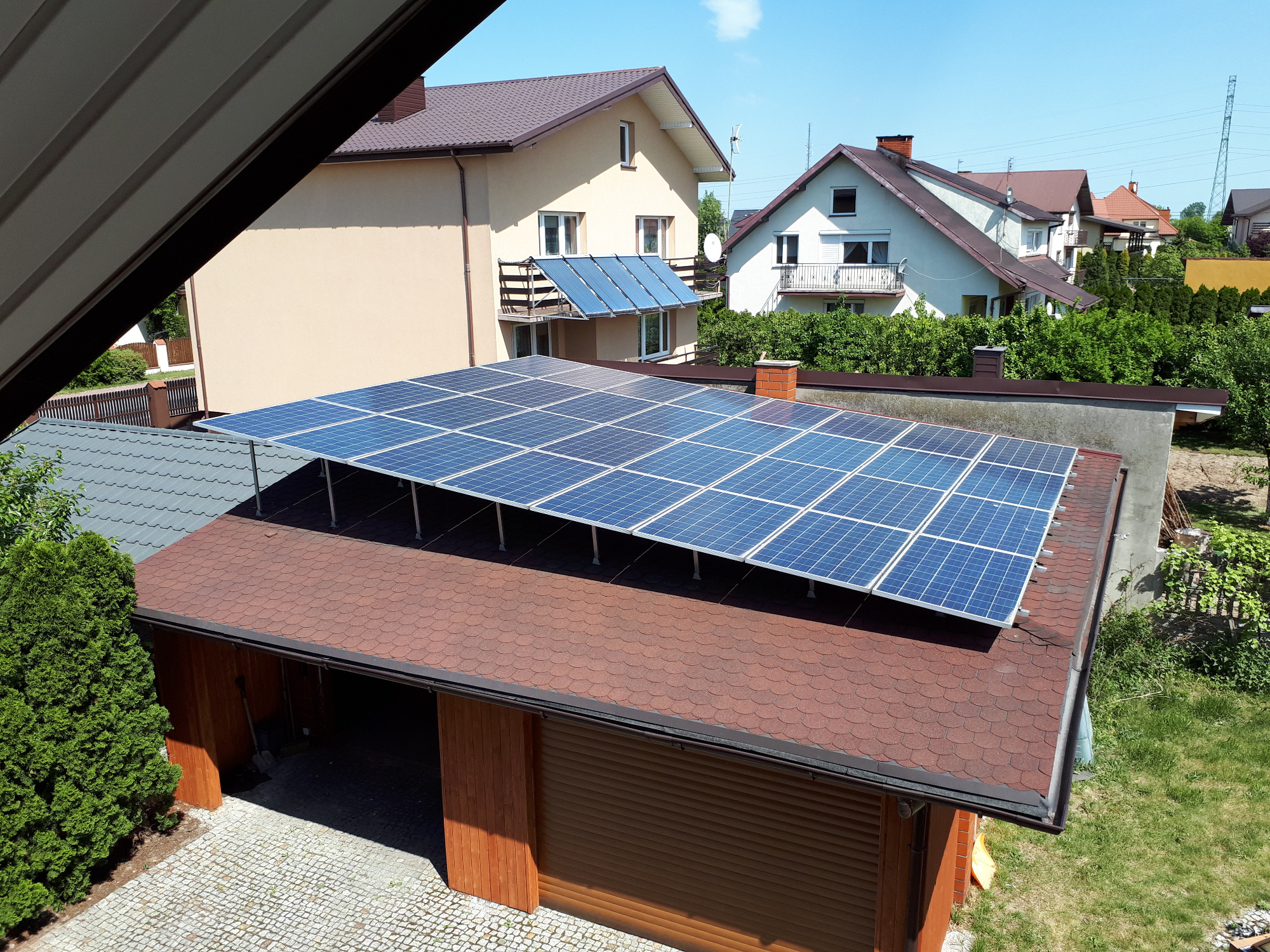Instalacja paneli słonecznych na dachu garażu z drewnianą bramą, w tle domy jednorodzinne i błękitne niebo.