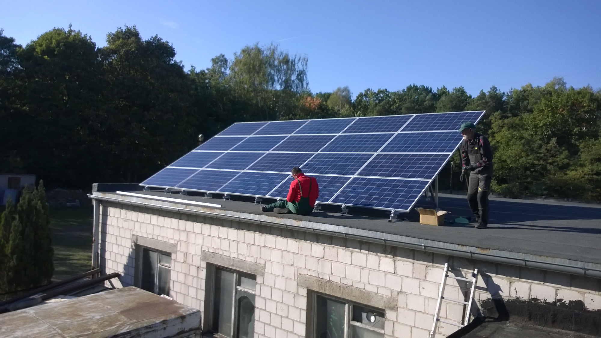Montaż paneli słonecznych na dachu budynku z białej cegły. Jeden pracownik siedzi na dachu, drugi stoi obok paneli, obok karton, drabina oparta o ścianę.