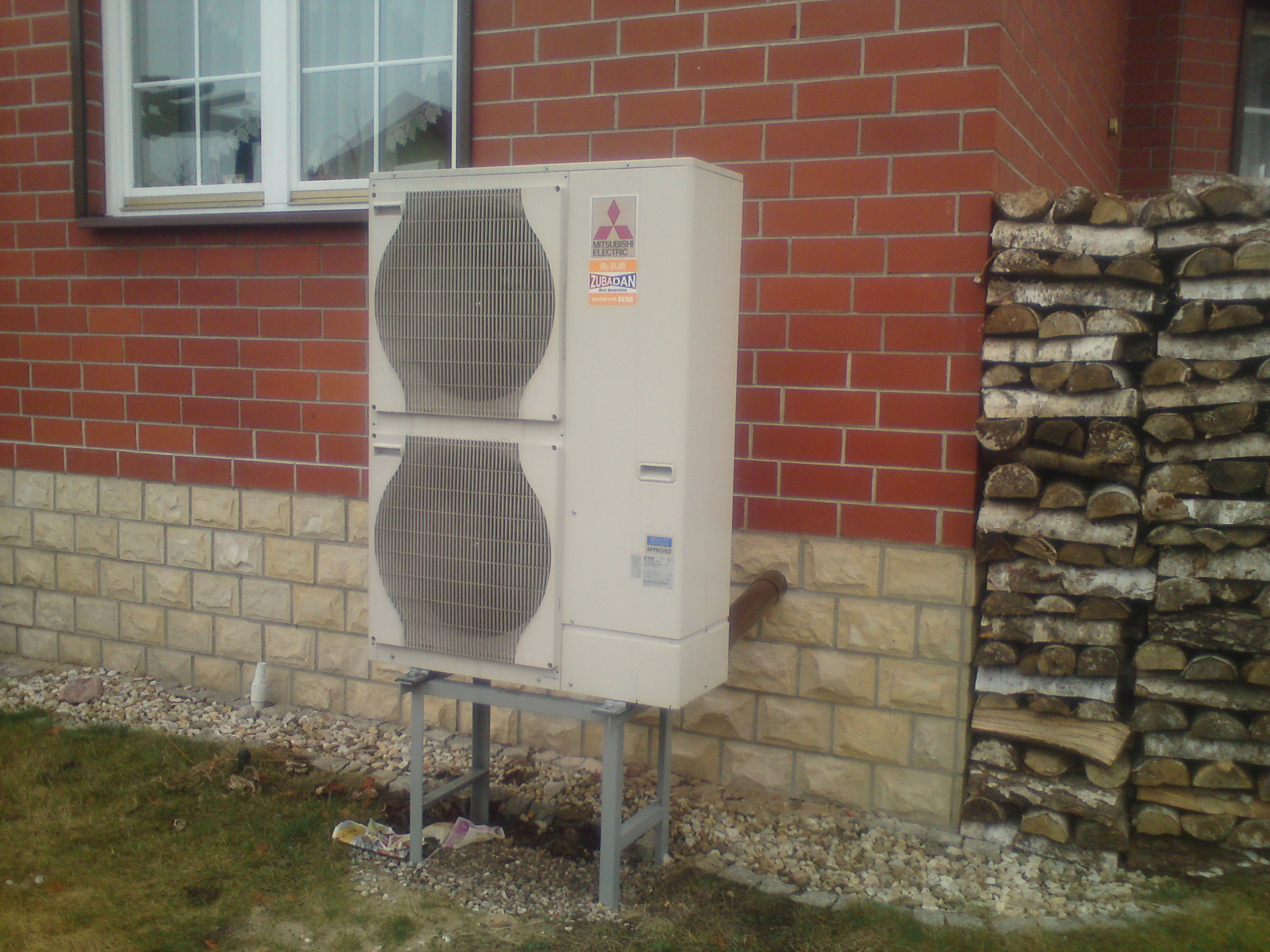 Biała zewnętrzna jednostka pompy ciepła Mitsubishi Zubadan, zamontowana na metalowym stelażu przy ścianie z czerwonej cegły, obok stosu drewna opałowego.