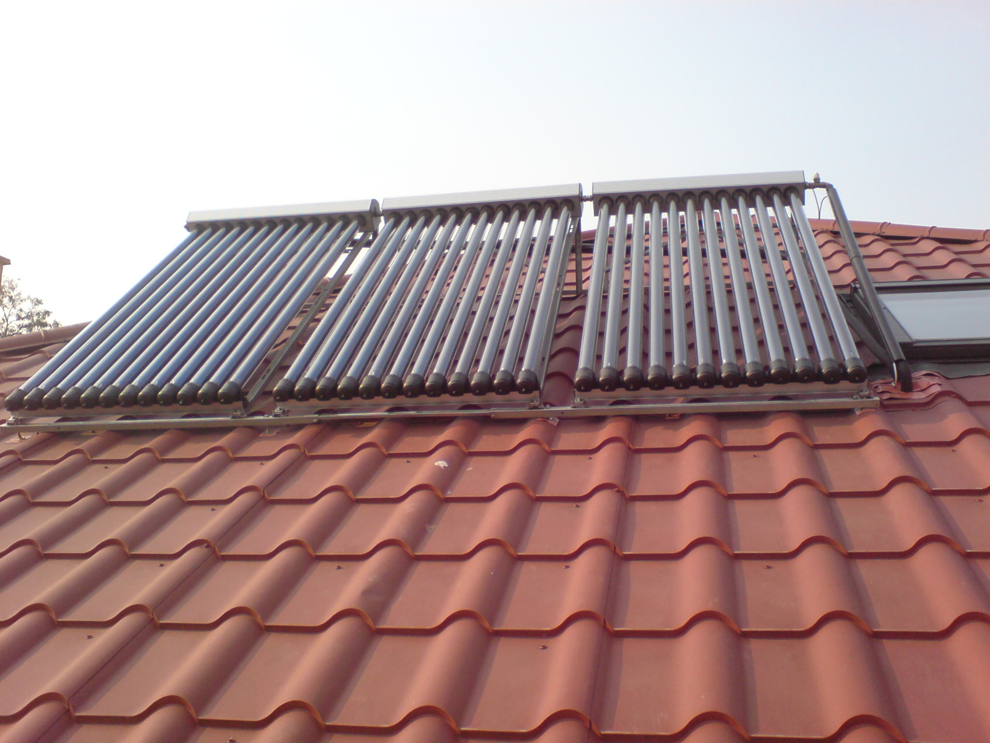 Dwa panele kolektorów słonecznych zamontowane na dachu z czerwonej dachówki, widoczny również fragment okna dachowego.