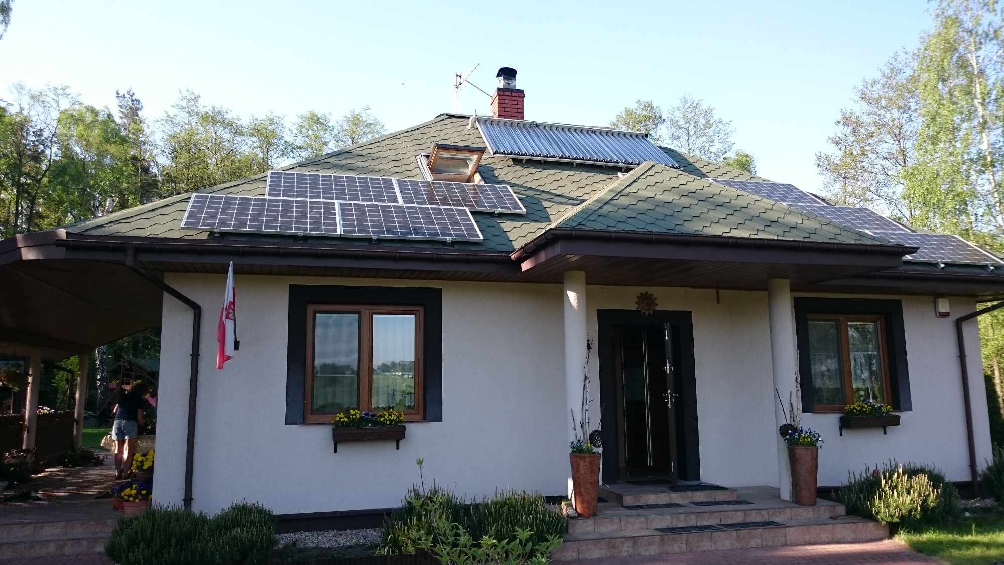 Dom jednorodzinny z panelami słonecznymi i kolektorami słonecznymi na dachu pokrytym zieloną dachówką, widoczne okno dachowe, flaga Polski, kwiaty w oknach i donicach.