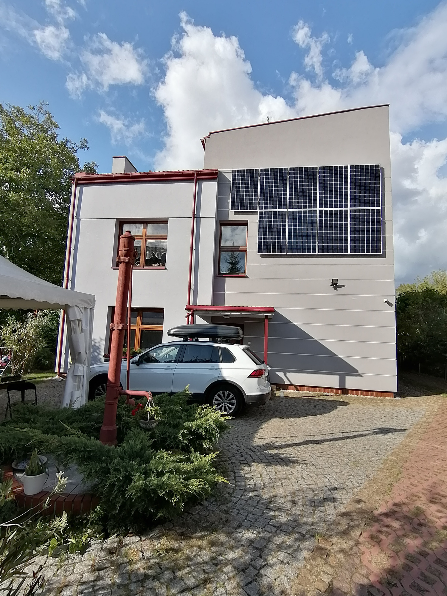 Nowoczesny dom z szarymi ścianami i panelami słonecznymi na elewacji, biały samochód z bagażnikiem dachowym zaparkowany przed budynkiem, stara czerwona pompa wodna na pierwszym planie.