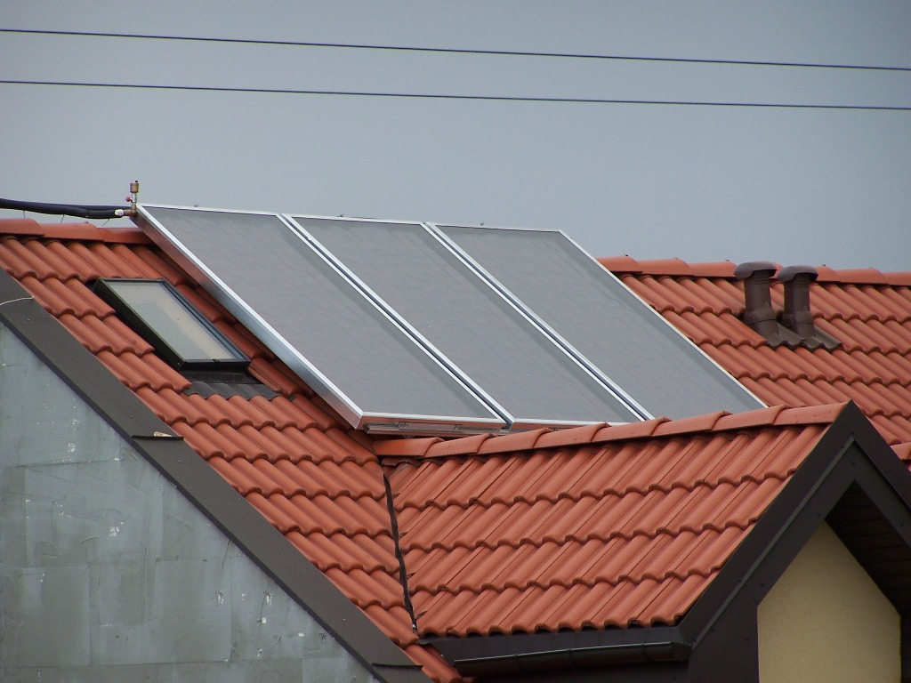 Trzy panele słoneczne z srebrną ramą zamontowane na czerwonym dachu z dachówki, obok okna dachowego i dwóch kominów wentylacyjnych, widoczne przewody i linia energetyczna na szarym tle nieba.