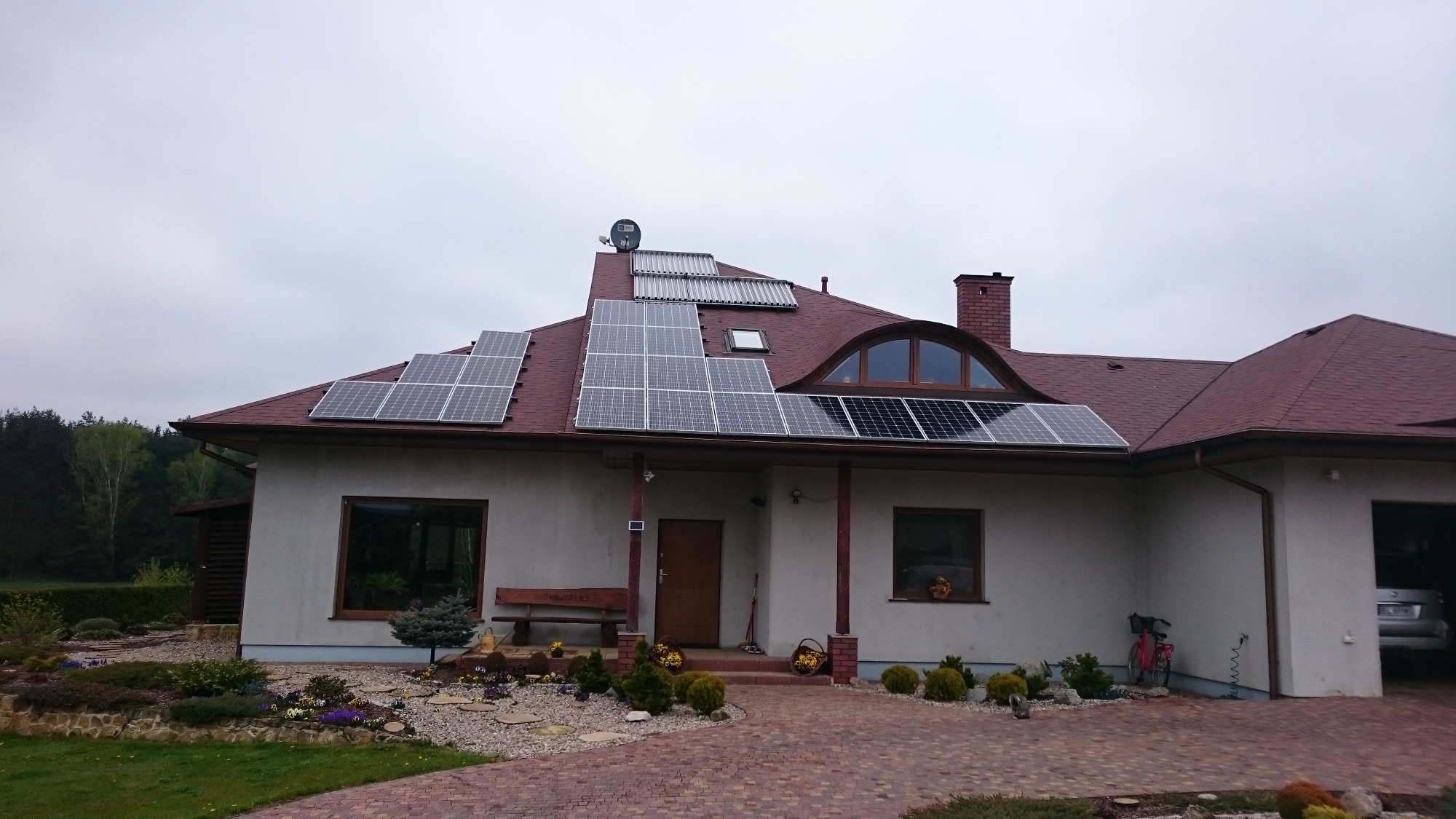 Dom jednorodzinny z panelami słonecznymi na dachu pokrytym czerwoną dachówką bitumiczną, dodatkowo kolektory słoneczne i satelita na dachu, brukowany podjazd i zadbany ogród.