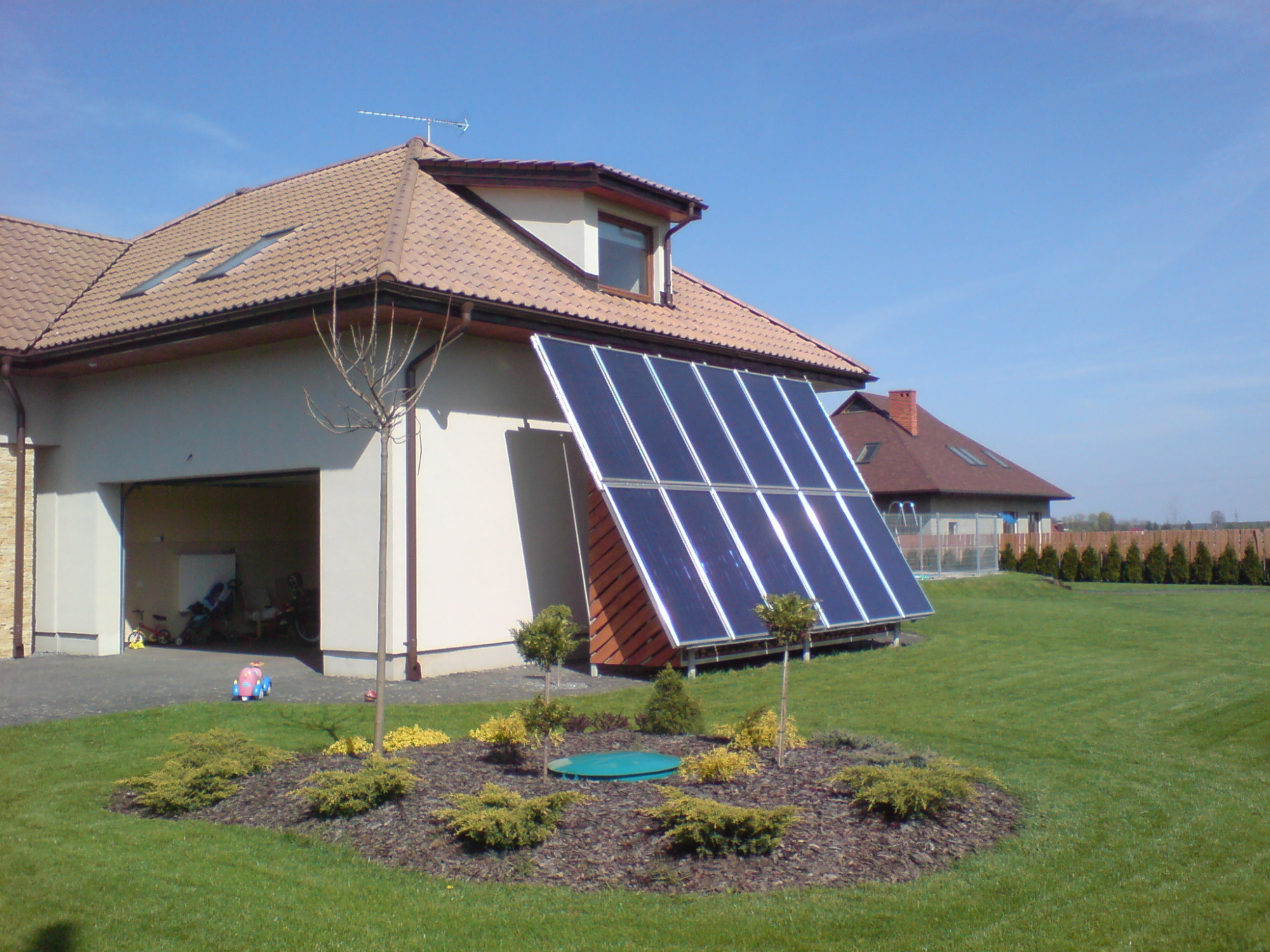 Panele słoneczne ustawione na trawniku obok domu z otwartym garażem, na pierwszym planie ozdobny klomb z niskimi krzewami i niebieską pokrywą studzienki.