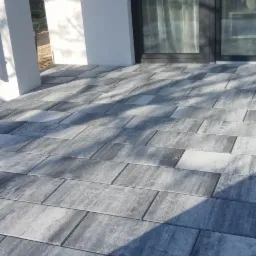 Szara, prostokątna kostka na tarasie przed nowoczesnym domem z dużymi oknami, widoczne cienie drzew.