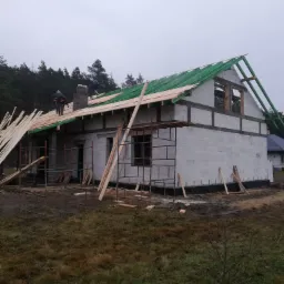 Budowa domu jednorodzinnego: surowy stan z zieloną konstrukcją dachu i pracownikami na górze, widok z zewnątrz na tle lasu w pochmurny dzień.