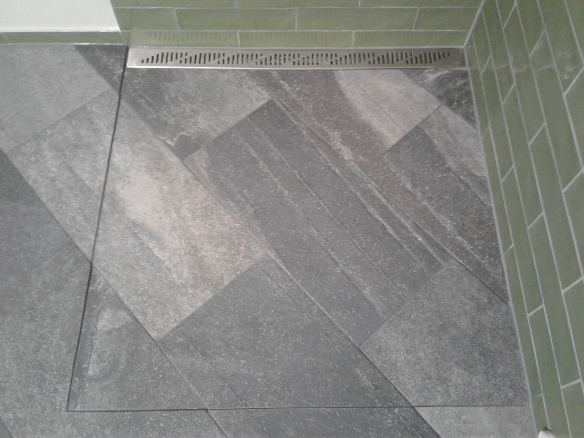 Ułożona szara podłoga z płytek w jodełkę, z odpływem liniowym i zielonymi płytkami ściennymi. Detal wykończenia łazienki.