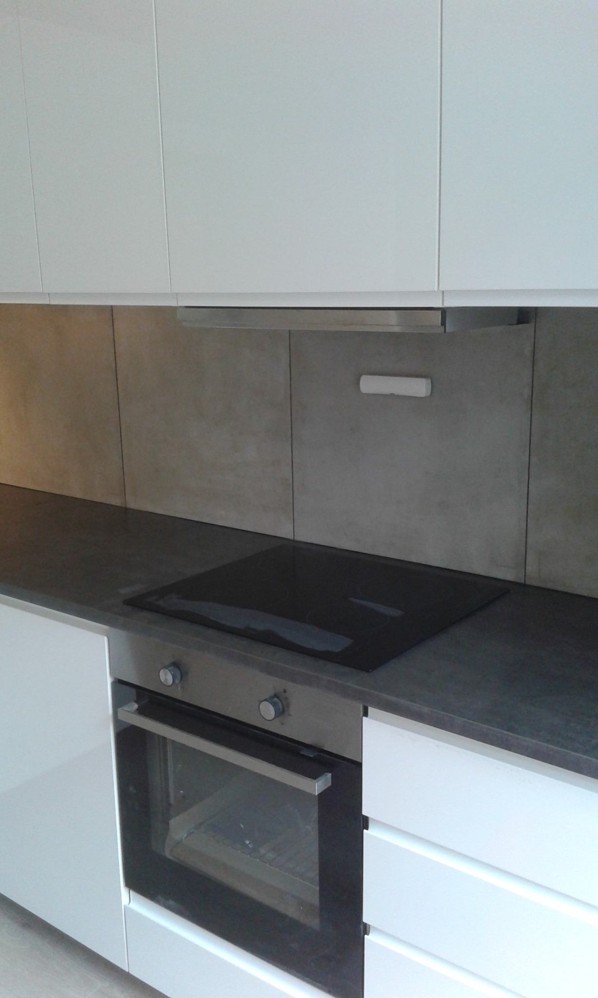 Nowoczesna kuchnia z białymi, lakierowanymi szafkami, szarym blatem i płytą indukcyjną. Minimalistyczny design, jasne płytki na ścianie, okap.