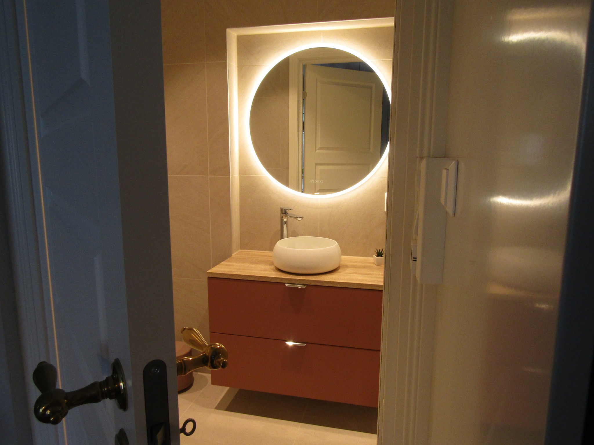 Nowoczesna łazienka z okrągłym lustrem LED, umywalką nablatową i szafką w kolorze terakoty. Ujęcie przez uchylone drzwi, widoczne detale wykończenia.