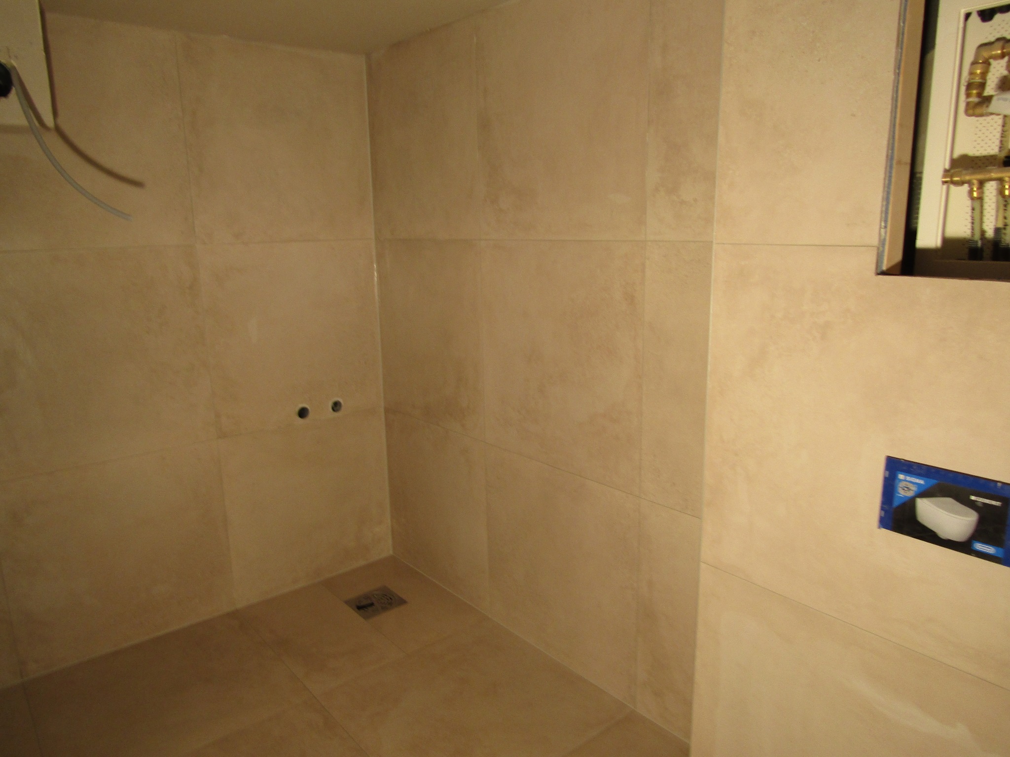 Wykończona łazienka z beżowymi płytkami, widoczny odpływ liniowy, instalacja podtynkowa WC i wyprowadzone rury. Minimalistyczny design.