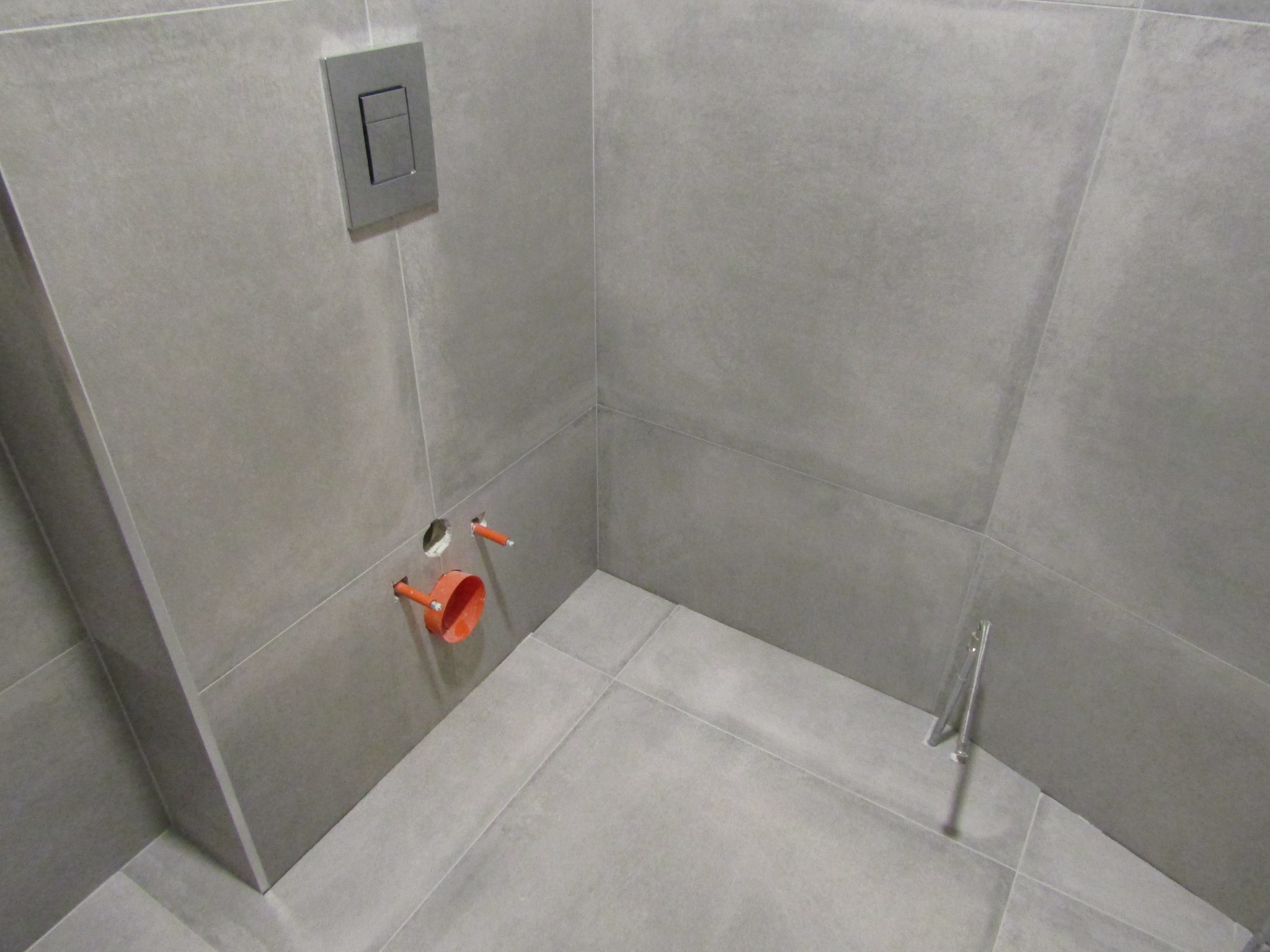 Szara łazienka w trakcie wykańczania: widoczne płytki, przycisk spłuczki, rury i wspornik. Minimalistyczny design, surowe wykończenie.