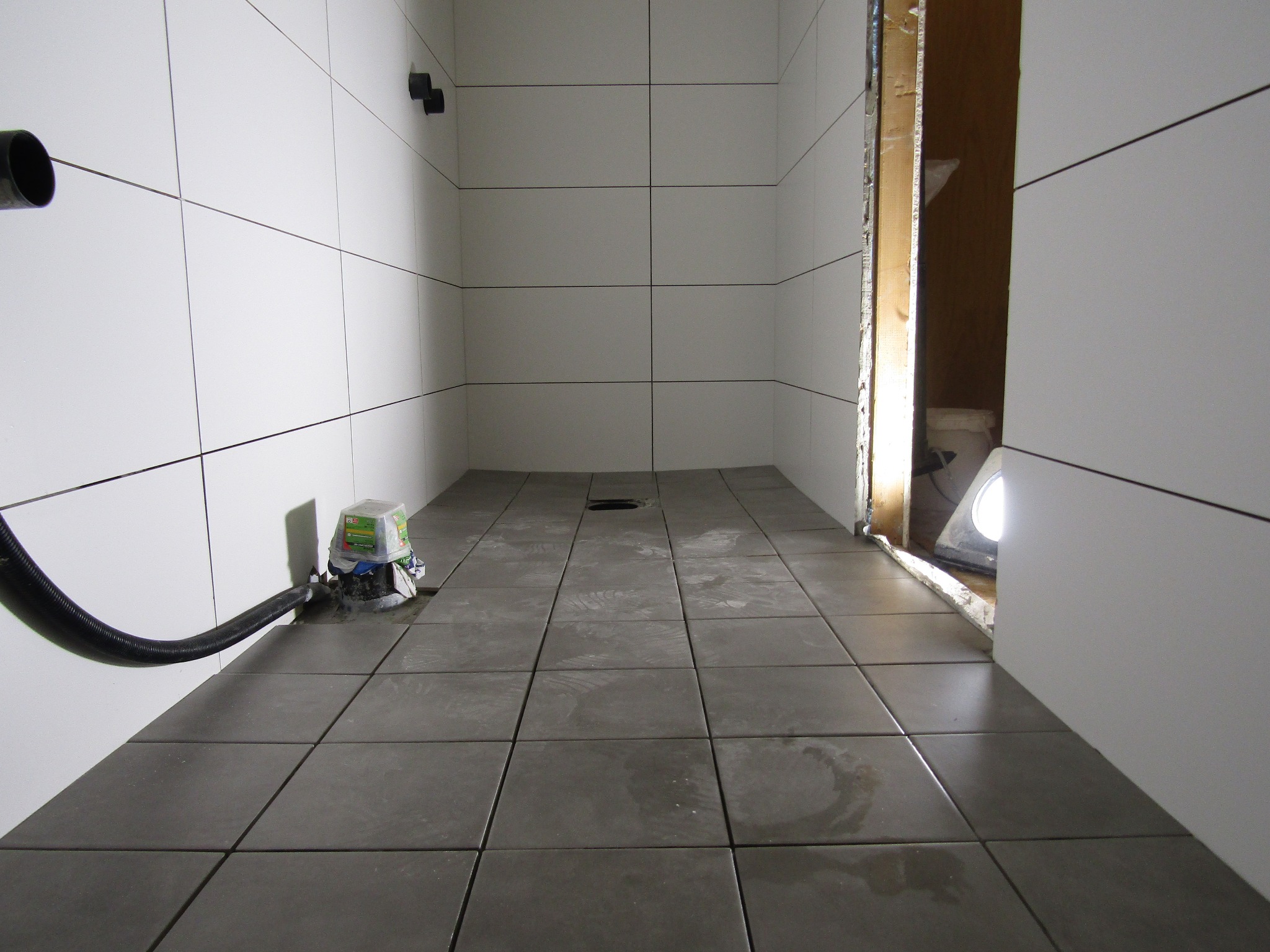 Nowoczesna kabina prysznicowa w trakcie wykańczania: białe płytki ścienne, szare płytki podłogowe, odpływ liniowy i widoczny element instalacji hydraulicznej.