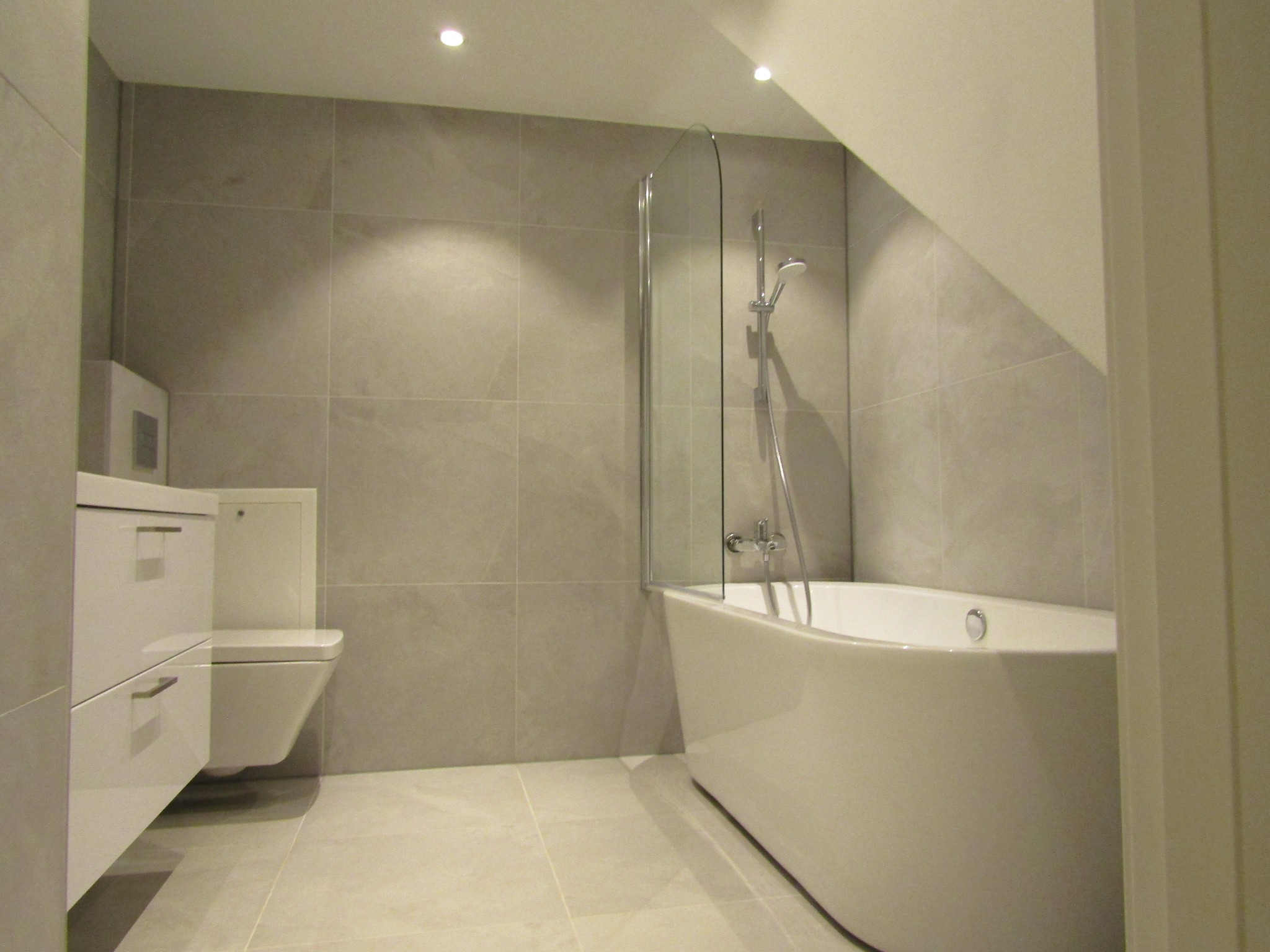 Jasna łazienka na poddaszu z wanną, szklanym parawanem i podwieszaną toaletą z szafką, wykończona płytkami w odcieniach szarości.