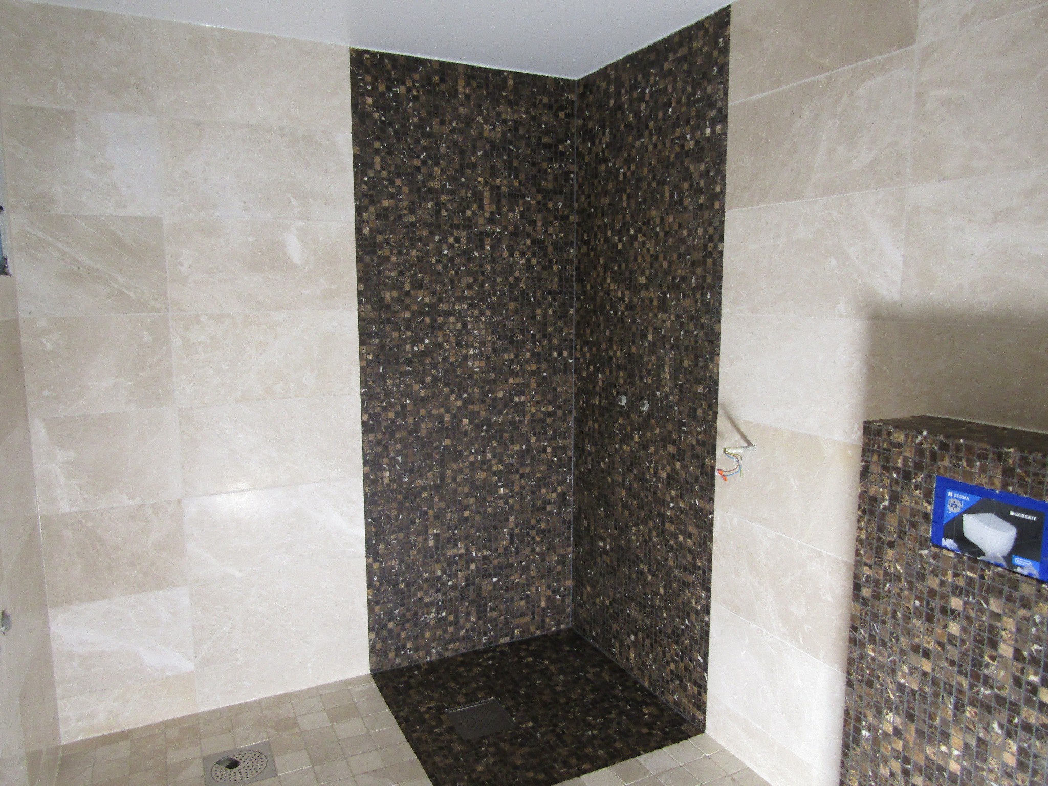 Wykończony brodzik z mozaiki w odcieniach brązu, z odpływem liniowym. Ściany wyłożone jasnymi, marmurkowymi płytkami. Widoczna instalacja podtynkowa.