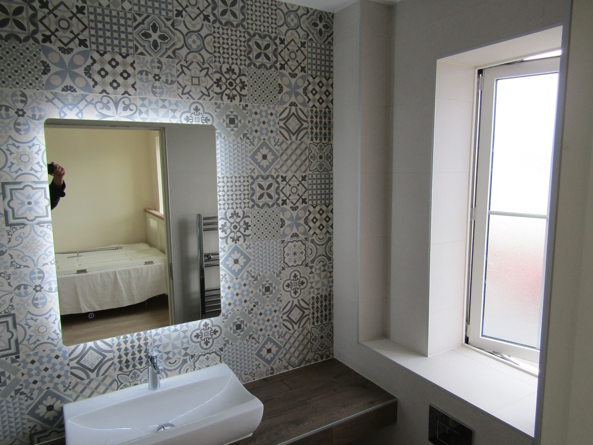 Wykończona łazienka z umywalką, lustrem w ozdobnych płytkach i oknem. Płytki ścienne w odcieniach szarości i błękitu z różnorodnymi wzorami. Widoczny fragment sypialni w lustrze.