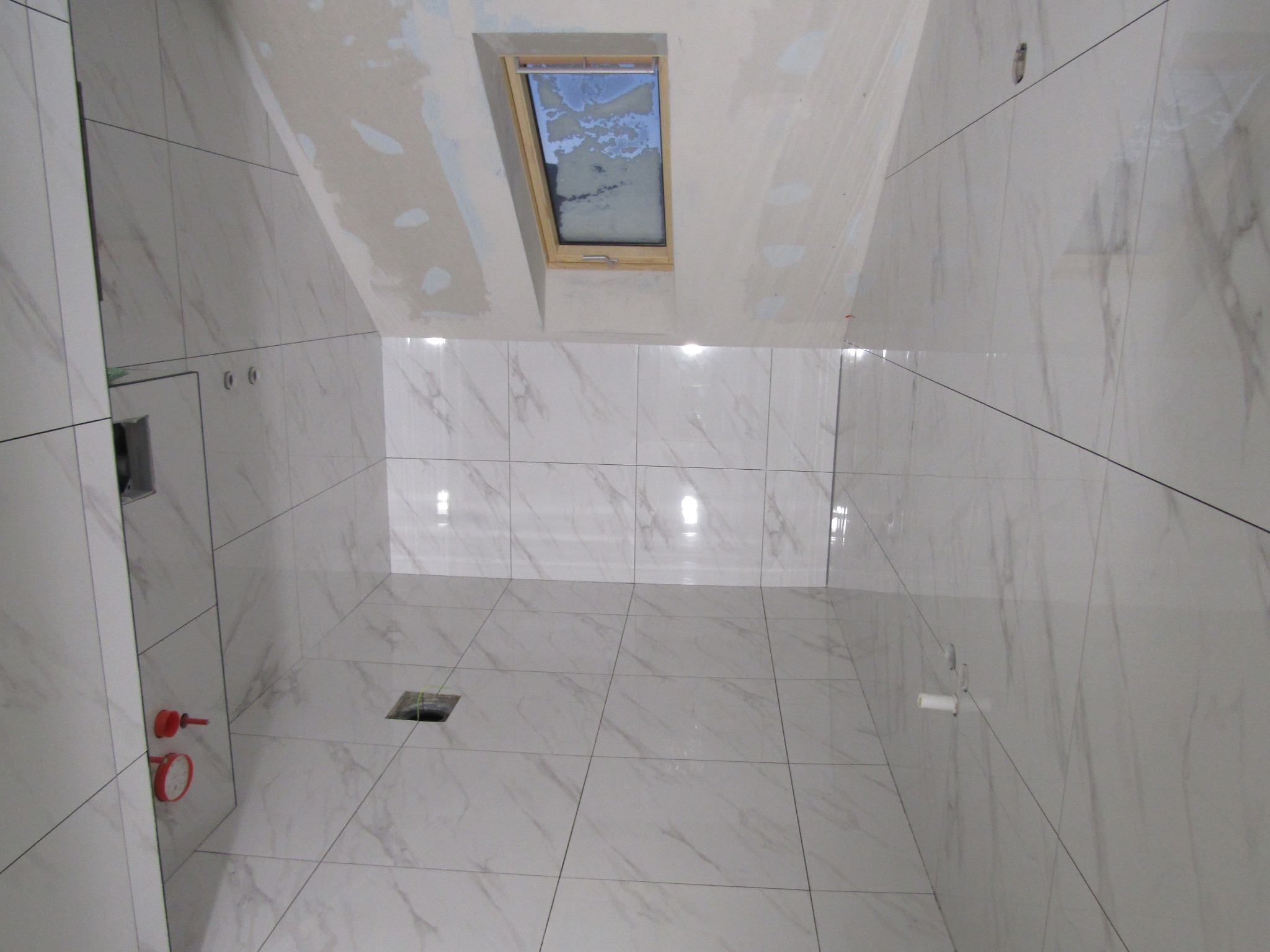 Wykończona łazienka na poddaszu z jasnymi, marmurkowymi płytkami, drewnianym oknem dachowym i przygotowaną instalacją wodno-kanalizacyjną.