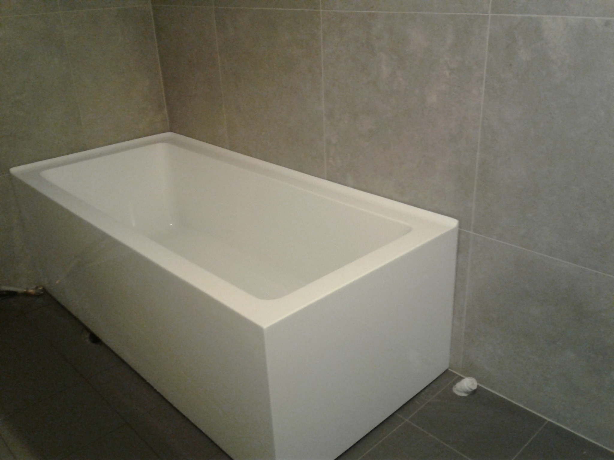 Biała, prostokątna wanna obudowana płytkami w szarej łazience. Widoczny dolny zawór odpływowy. Minimalistyczny design i świeżo wykończone wnętrze.