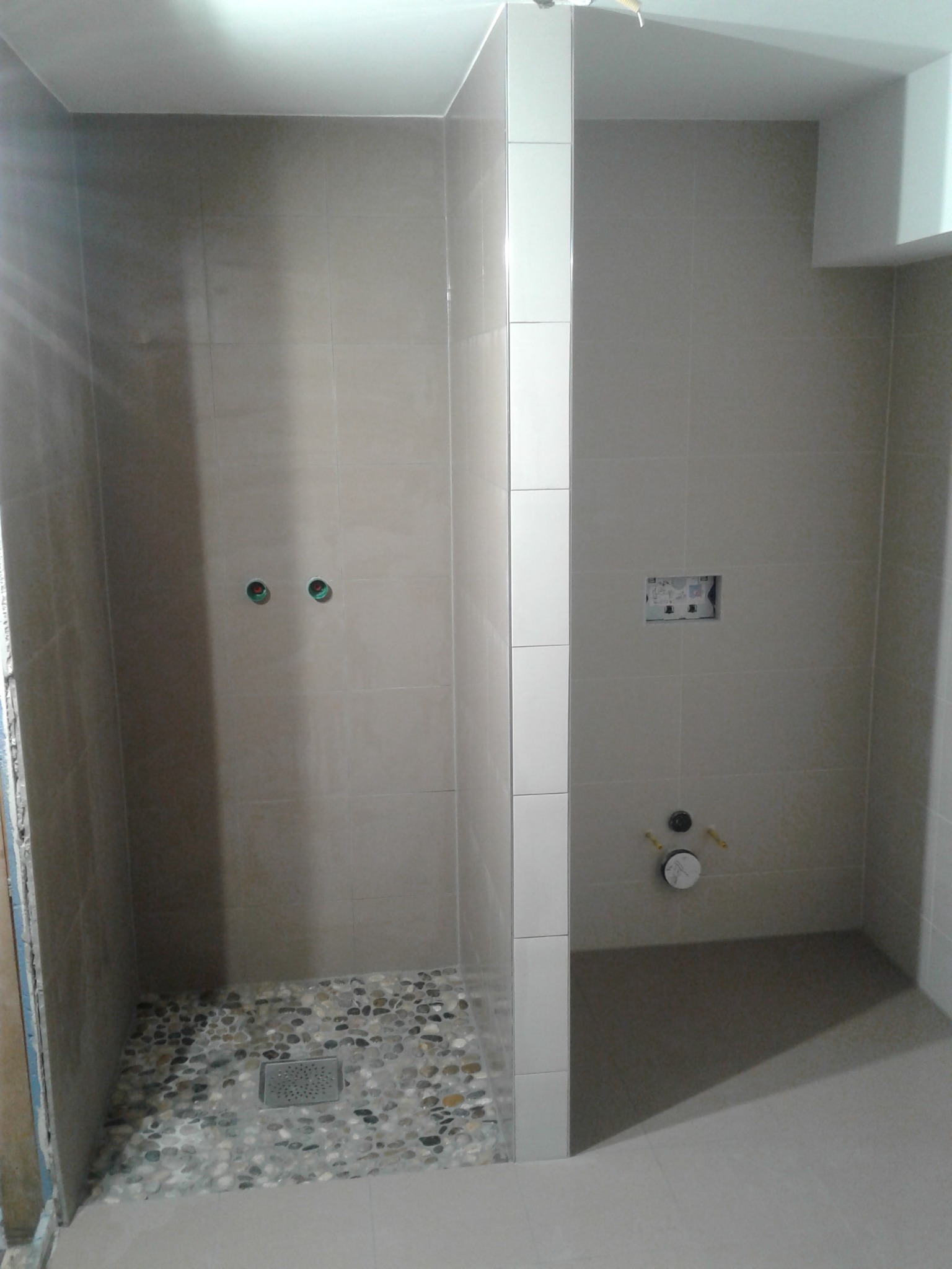 Wykończona kabina prysznicowa z beżowymi płytkami i kamienną mozaiką na podłodze. Widoczne odpływ liniowy i przygotowane punkty instalacyjne.