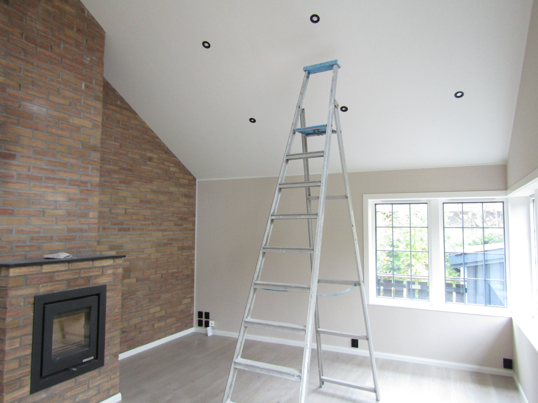 Wnętrze po remoncie: ściana z cegły przy kominku, świeżo pomalowane ściany w odcieniach beżu, drabina oparta o ścianę, nowe oświetlenie sufitowe, okno z widokiem na zieleń.