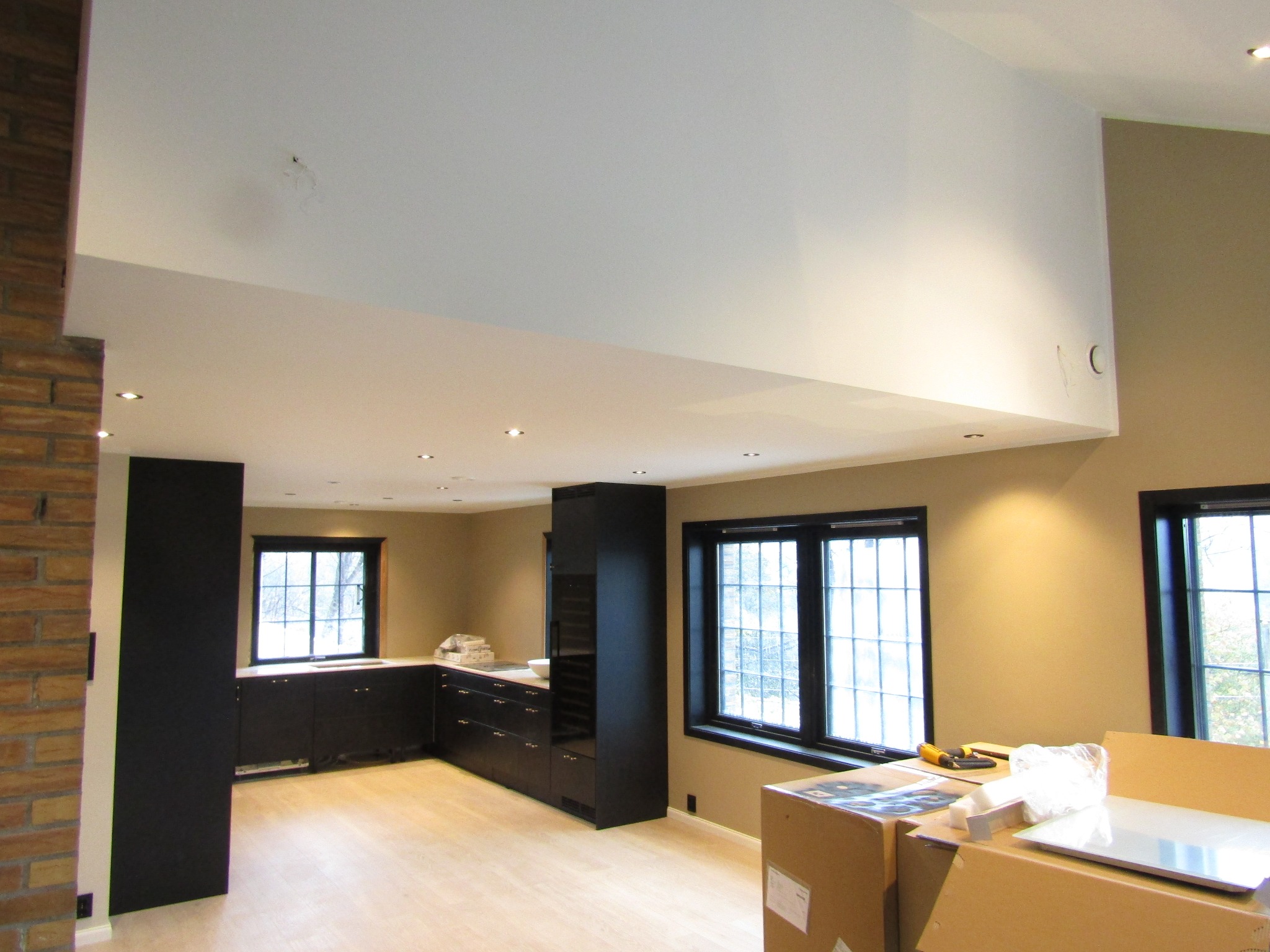 Jasne wnętrze z czarnymi meblami kuchennymi i dużymi oknami. Na pierwszym planie kartony z elementami wyposażenia. Widoczny fragment ściany z cegły.