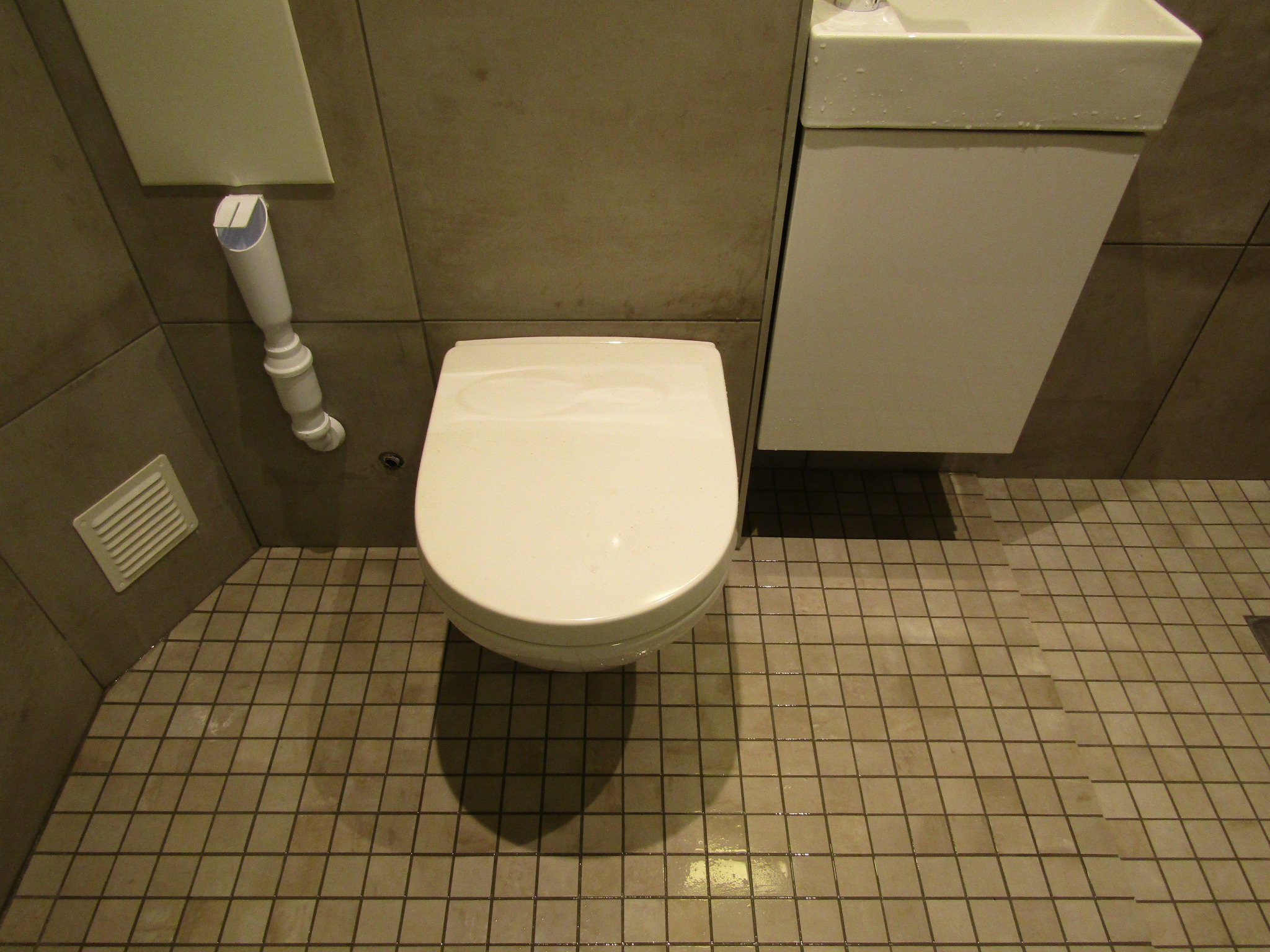 Jasna toaleta z deską, umywalka i brązowe płytki podłogowe w nowoczesnej łazience. Widoczny fragment wentylacji i rury. Minimalistyczne wykończenie.