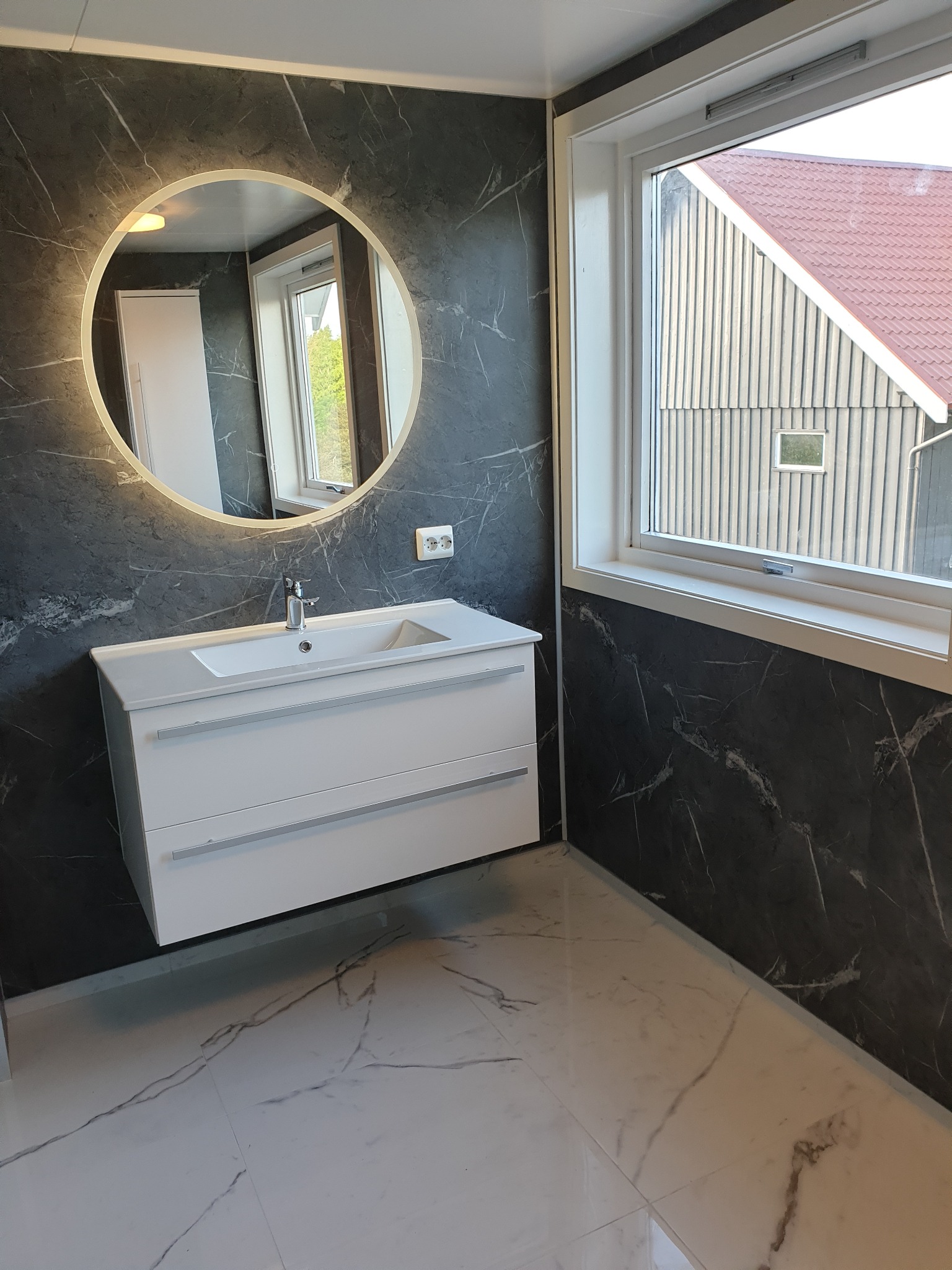 Nowoczesna łazienka z umywalką na szafce, okrągłym lustrem z podświetleniem LED i płytkami imitującymi marmur w odcieniach szarości i bieli. Widok na okno.