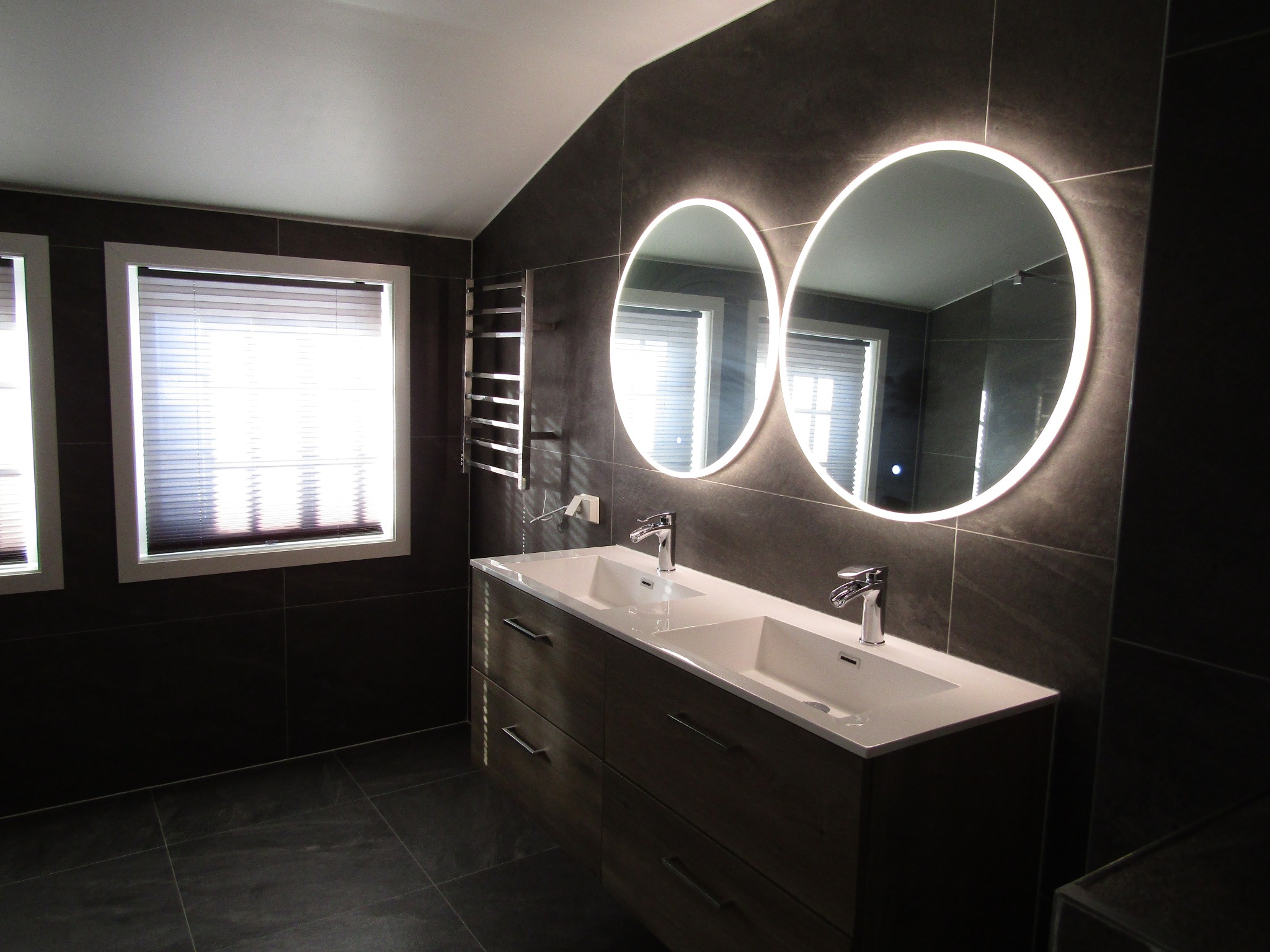Ciemna, elegancka łazienka z dwoma okrągłymi lustrami z podświetleniem LED, umywalką nablatową i szarą glazurą. Grzejnik drabinkowy i żaluzje w oknach dopełniają wystrój.