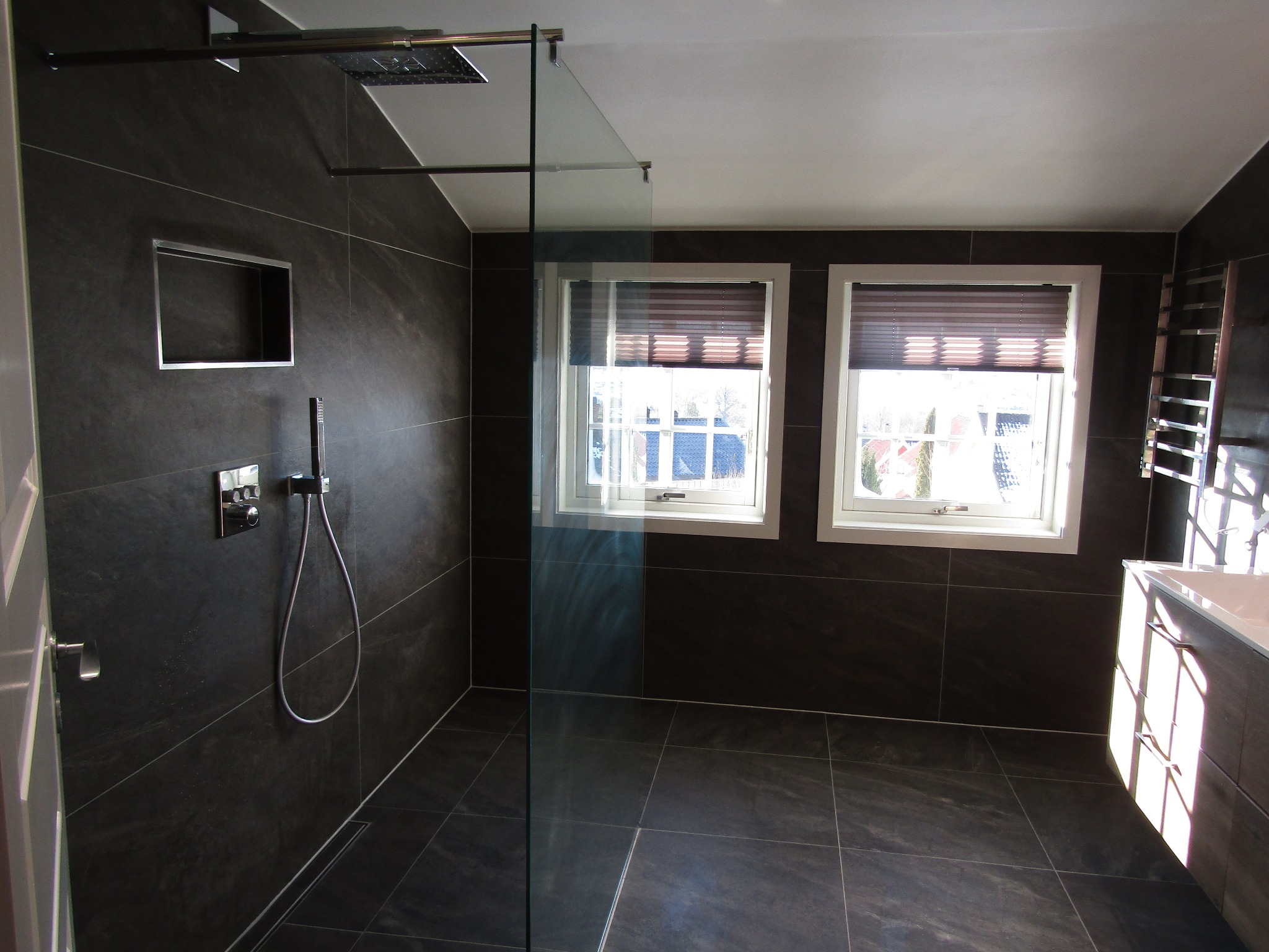 Nowoczesna łazienka z ciemnymi płytkami, kabiną prysznicową typu walk-in i oknami z żaluzjami. Widoczna wnęka w ścianie i bateria prysznicowa.