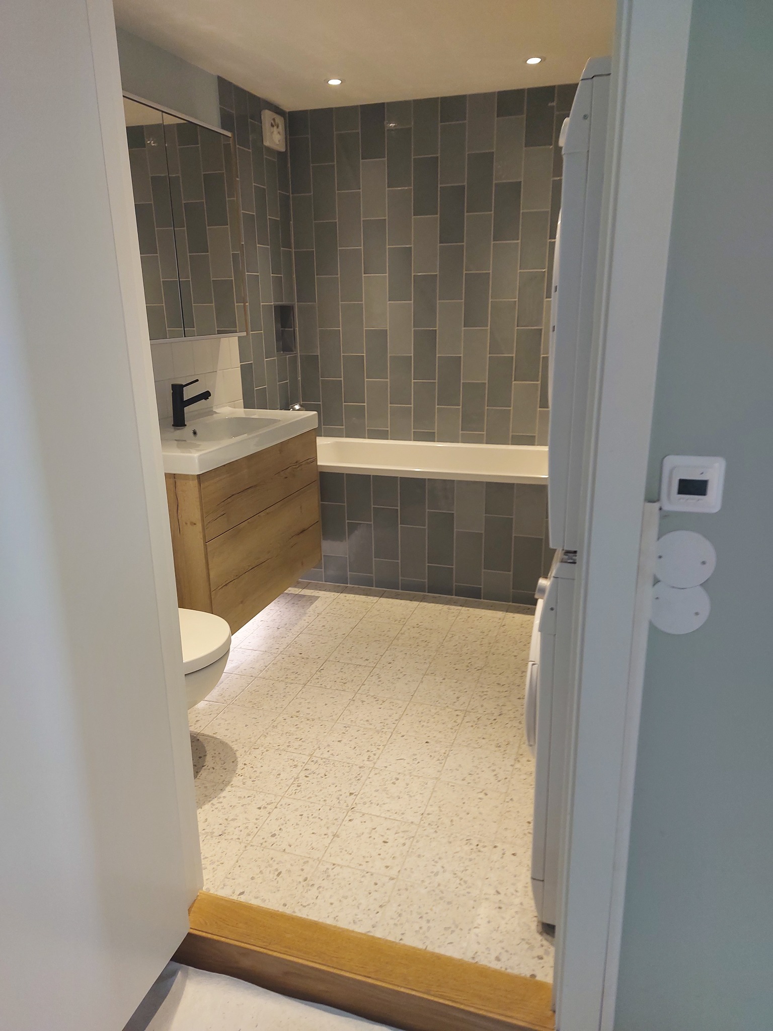 Nowoczesna łazienka z drewnianą szafką pod umywalką, szarą glazurą w cegiełkę i beżową podłogą z terazzo, widziana zza uchylonych białych drzwi.