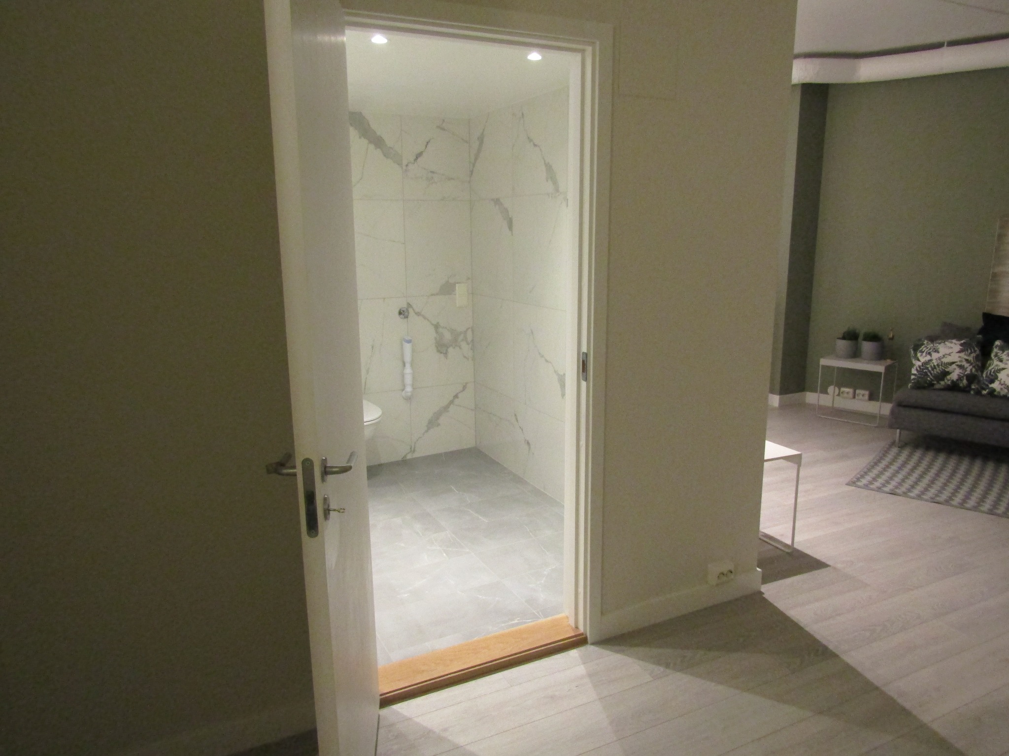 Wykończona łazienka z marmurowymi płytkami, widoczna przez otwarte drzwi. Minimalistyczny styl, szare płytki podłogowe, toaleta w lewym rogu. W tle salon z sofą.