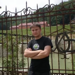 Mężczyzna stoi przed ozdobnym, rdzewiejącym ogrodzeniem z kutego żelaza na tle zielonego wzgórza z budynkiem.