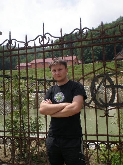 Mężczyzna stoi przed ozdobnym, rdzewiejącym ogrodzeniem z kutego żelaza na tle zielonego wzgórza z budynkiem.