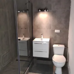 Nowoczesna łazienka w szarościach z kabiną prysznicową typu walk-in z odpływem liniowym, wiszącą szafką z umywalką i toaletą.