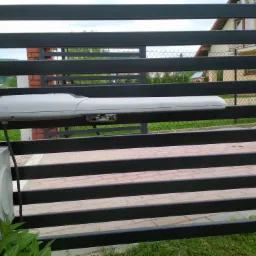 Zewnętrzny napęd do bramy przesuwnej z czarnymi, poziomymi szczeblami, zamontowany na ceglanym słupku z widoczną skrzynką sterującą i zielenią u podstawy.