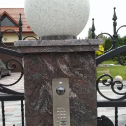 Zewnętrzny panel wideodomofonu z kamerą i klawiaturą numeryczną, zamontowany na marmurowym słupku bramy, zwieńczonym granitową kulą. W tle widoczny ogród i fragment domu.