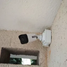 Kamera monitoringu BCS zamontowana na chropowatej, jasnej ścianie, widok z dołu, w tle widoczne drzewa przez otwór w murze.