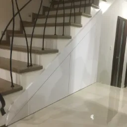 Białe szafki w zabudowie pod schodami z drewnianymi stopniami i czarną, dekoracyjną balustradą, widoczne drzwi w tle.