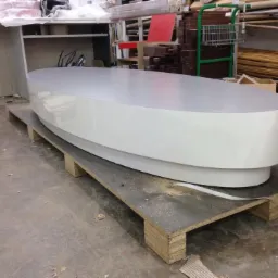 Owalny, biały blat drewniany z połyskiem, umieszczony na palecie w warsztacie stolarskim, w tle widoczne półki z materiałami.