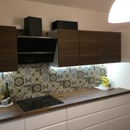 NETMAX Sp. z o.o. - Nowoczesna kuchnia z drewnianymi szafkami wiszącymi, białymi szafkami dolnymi z szufladami, blatem z drewna, dekoracyjnymi płytkami na ścianie i czarną płytą indukcyjną.