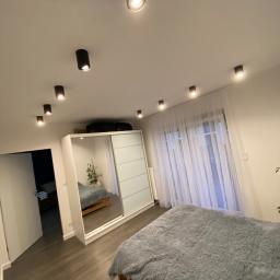 NETMAX Sp. z o.o. - Nowoczesna sypialnia z drewnianą podłogą, łóżkiem z szarym pluszowym pledem, białą szafą przesuwną z lustrem i rzędem czarnych punktowych lamp sufitowych.