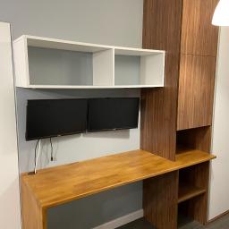 NETMAX Sp. z o.o. - Drewniane biurko z szafką i półkami, dwa monitory Dell przymocowane do ściany, biała szafka wisząca powyżej, szare ściany, nowoczesny design.