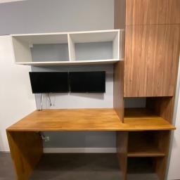 NETMAX Sp. z o.o. - Drewniane biurko na wymiar z szafką i półkami, dwa monitory na ścianie, białe półki wiszące, szare ściany.