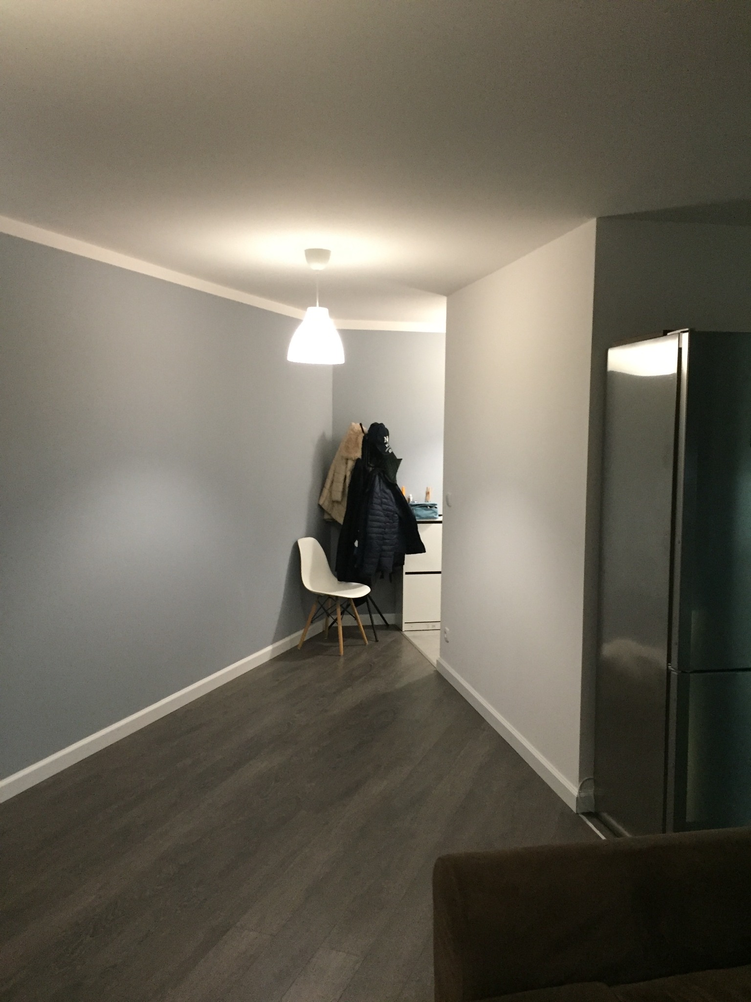 Wnętrze mieszkania po remoncie: szare ściany, ciemna podłoga, białe listwy przypodłogowe, widoczny fragment kuchni z białymi szafkami i lodówką, białe krzesło i wieszak na ubrania.