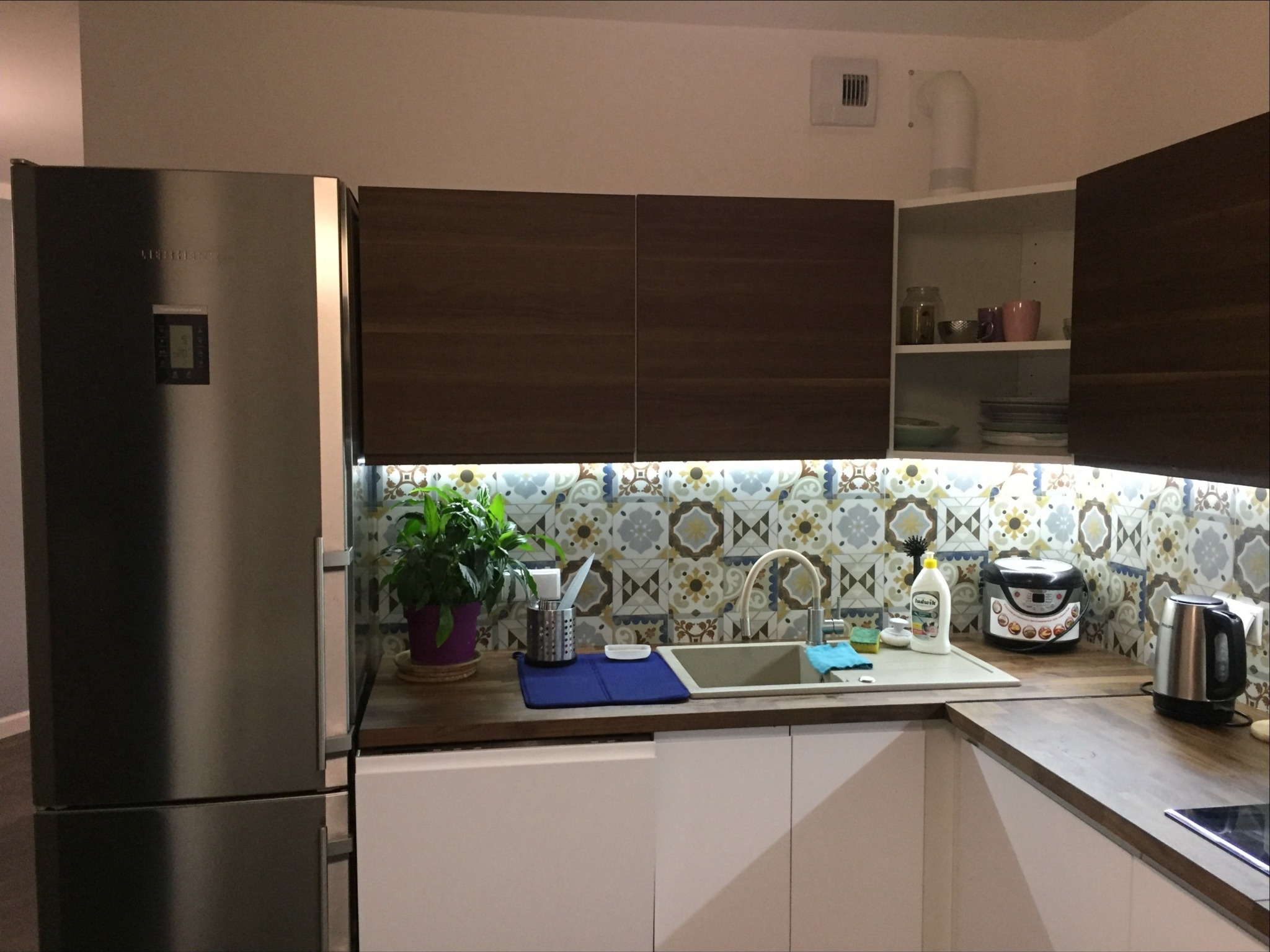 Nowoczesna kuchnia z białymi szafkami, drewnianymi blatami, mozaiką nad zlewem, i srebrną lodówką Liebherr po lewej stronie.