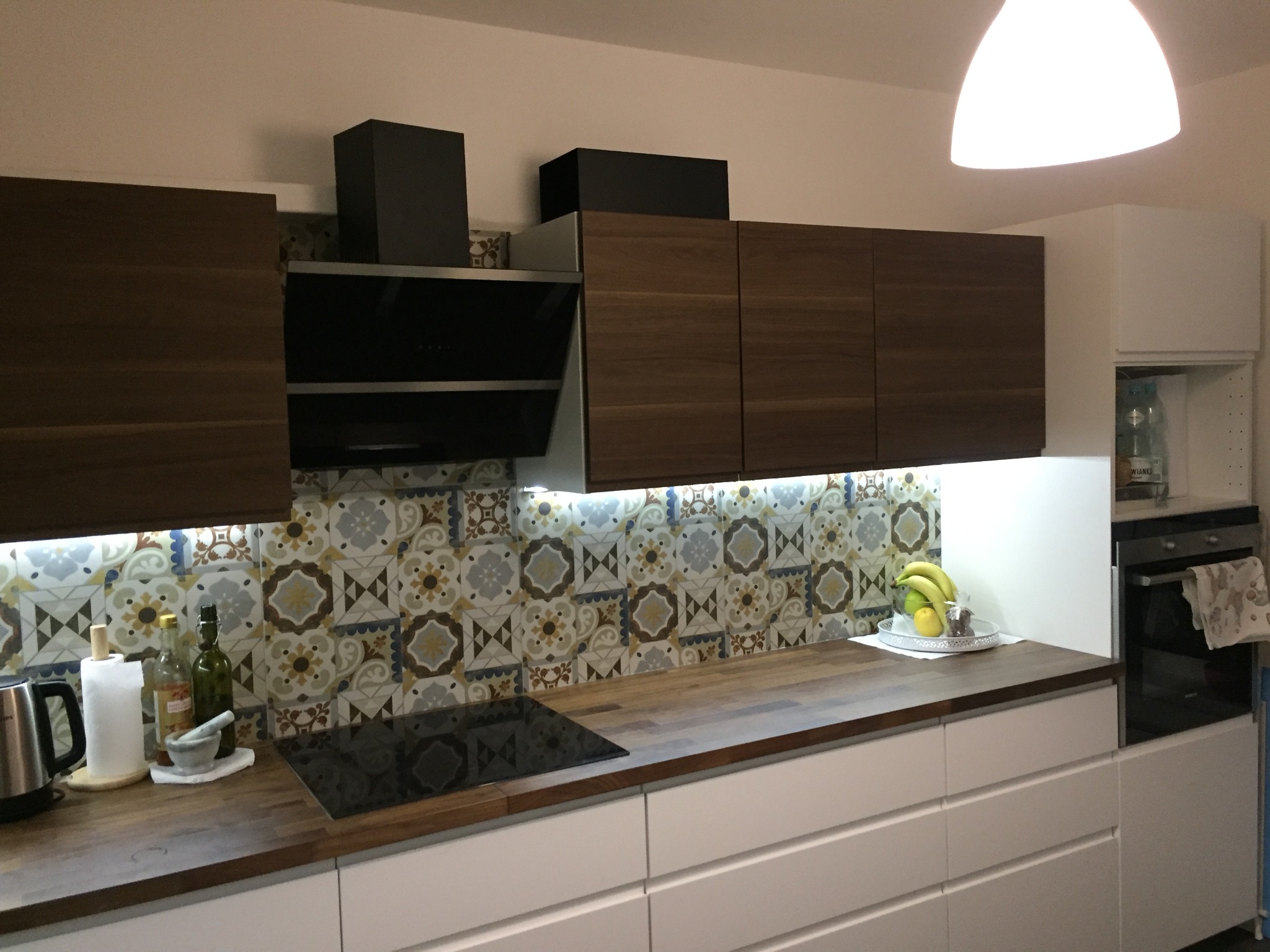 Nowoczesna kuchnia z drewnianymi szafkami wiszącymi, białymi szafkami dolnymi z szufladami, blatem z drewna, dekoracyjnymi płytkami na ścianie i czarną płytą indukcyjną.