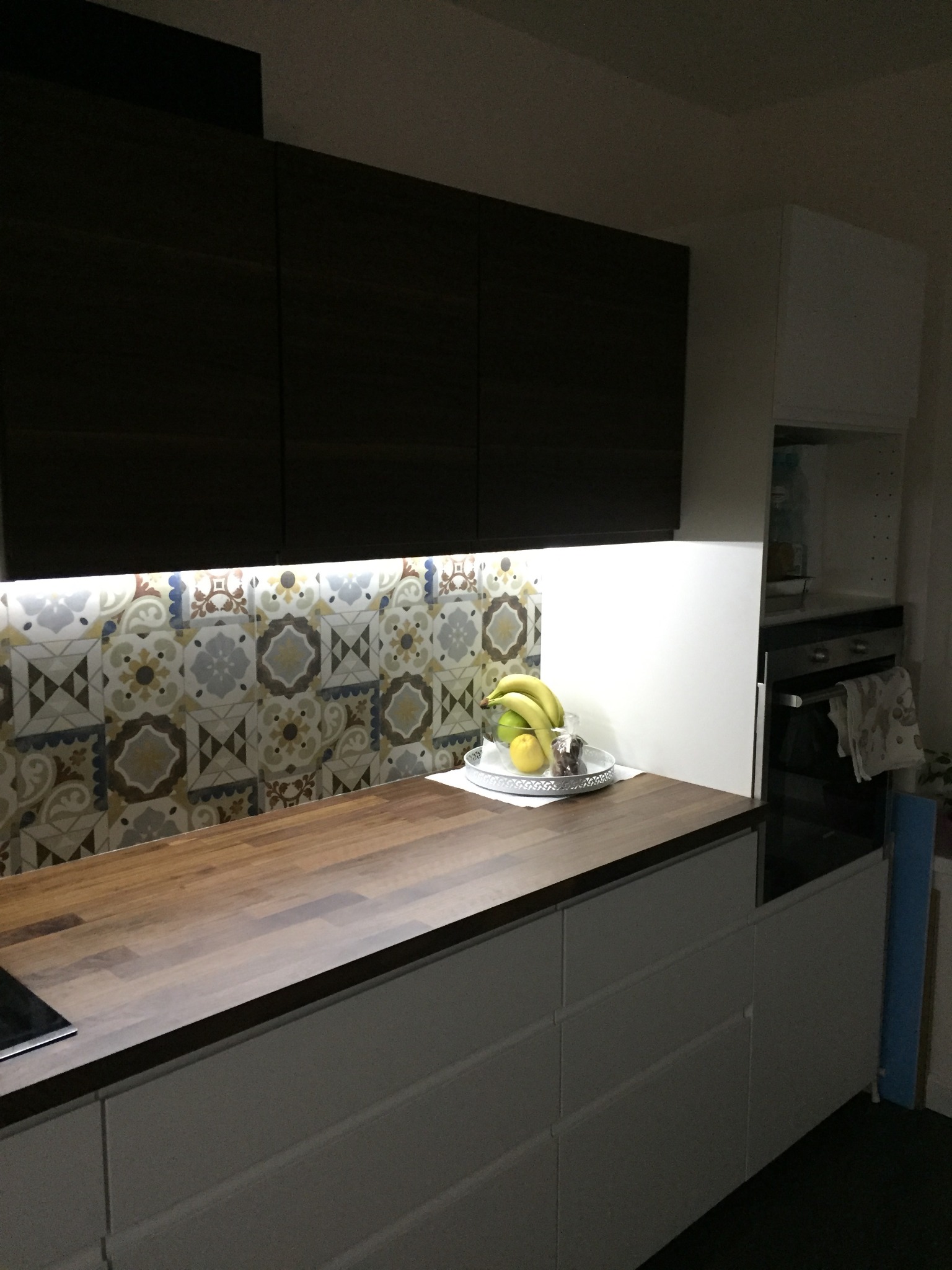 Nowoczesna kuchnia z ciemnymi górnymi szafkami, drewnianym blatem, wzorzystą ścianą nad blatem i owocami na srebrnej tacy.