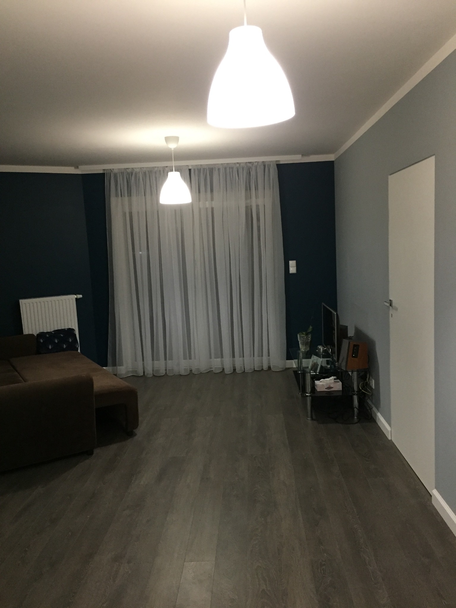 Nowoczesny salon z brązową sofą, drewnianą podłogą i oknem z białymi zasłonami, oświetlony dwoma lampami wiszącymi.