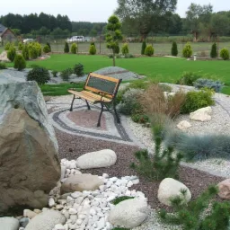 Ogród z ławką, dużym kamieniem i ozdobnymi ścieżkami z kostki granitowej oraz żwiru, w tle dom i równo przystrzyżony trawnik.