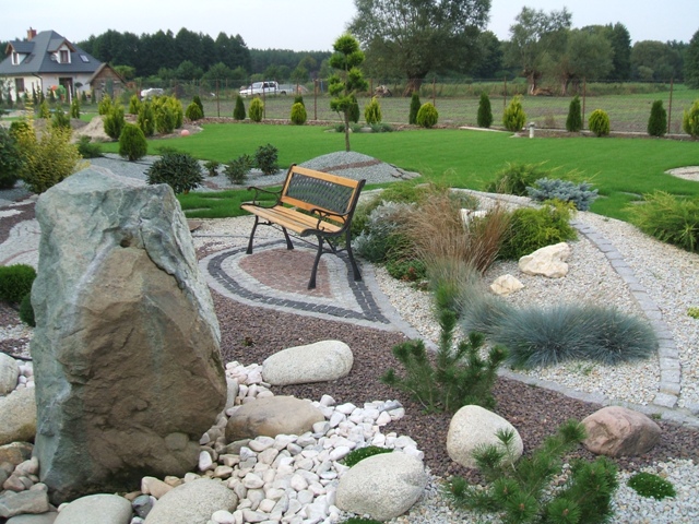 Ogród z ławką, dużym kamieniem i ozdobnymi ścieżkami z kostki granitowej oraz żwiru, w tle dom i równo przystrzyżony trawnik.